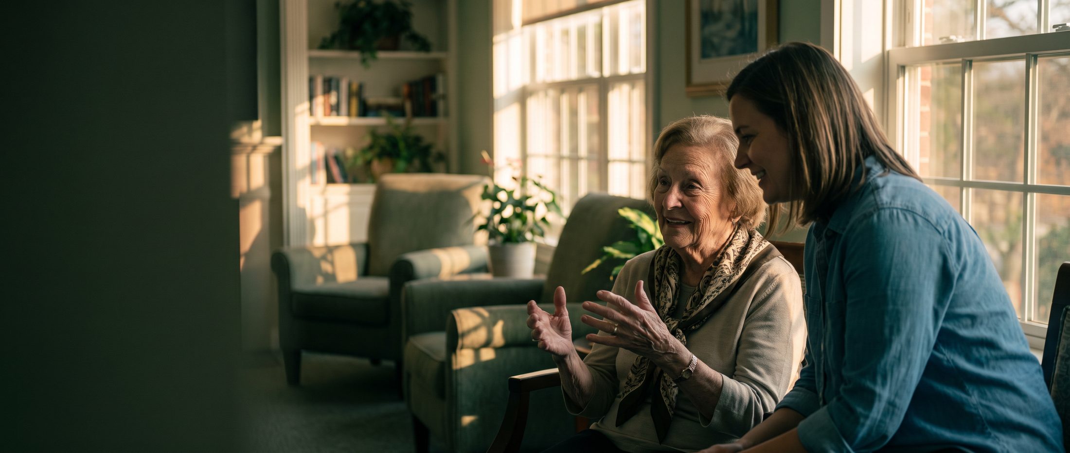 An older woman shares a story with a staff member in a sunlit senior living lounge.
